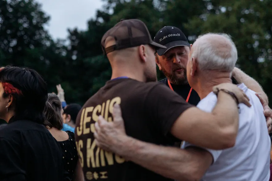 Three men praying with their arms around one another, one wearing a Jesus Is King shirt.
